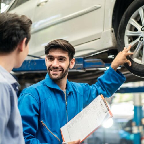 Vehicle maintenance handsome man servicer explain car condition, point at vehicle part to Asian customer in garage. Automotive mechanic hold automotive checklist document. Car repair service concept.